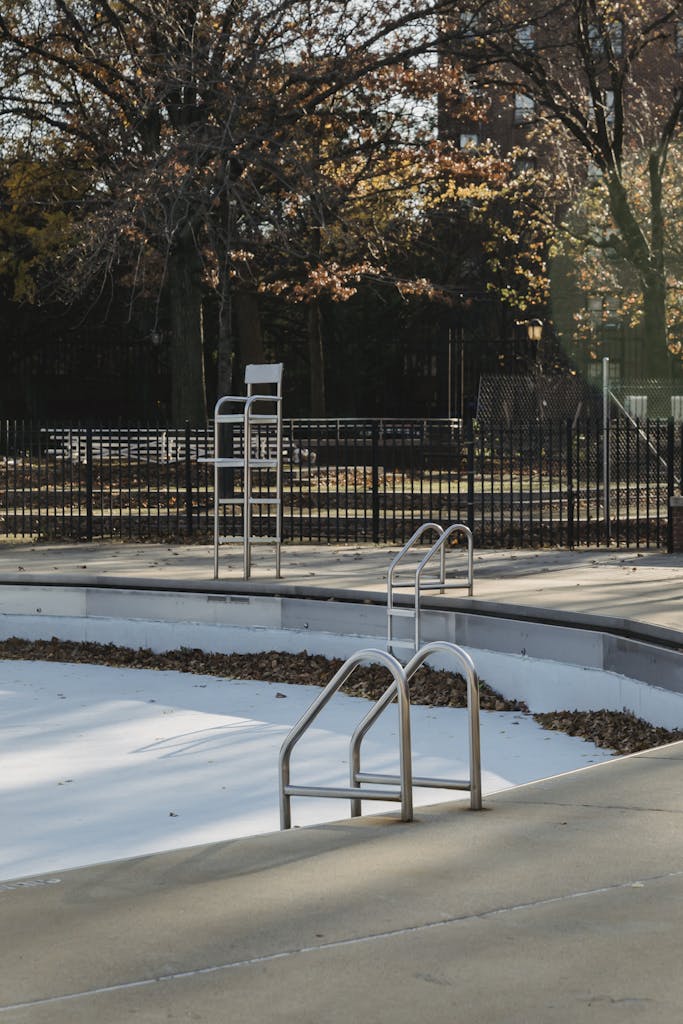 Empty waterless pool with metal ladders near tall chair in sunny autumn day near fence in park with trees and foliage in city street